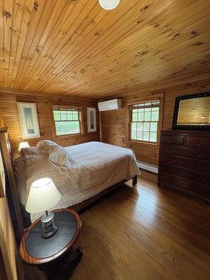 Une chambre à coucher dans une cabane en bois, avec un lit, des lampes de chevet, une commode et une fenêtre donnant sur un paysage verdoyant.