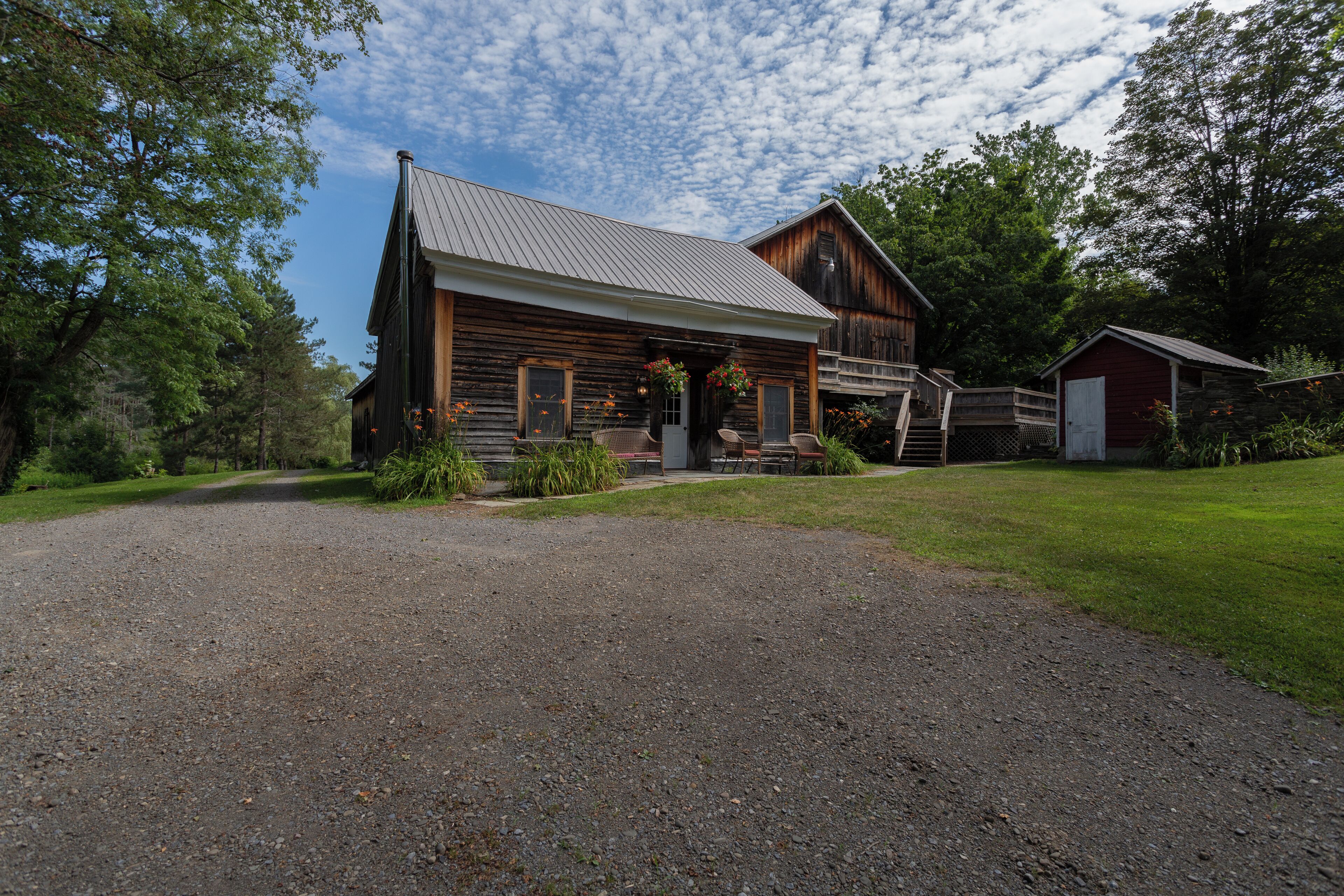 A rustic log cabin with a gravel driveway, a wooden porch, and a small adjacent shed.