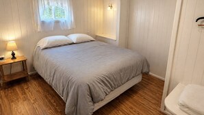 A bedroom with a bed, bedside table with lamp, wooden floor, and white walls.