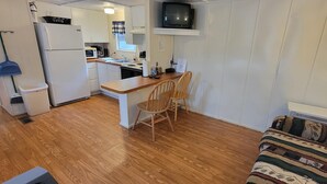 A compact kitchen with white cabinets, a microwave, a stove, and a sink. There is a wooden dining table with two chairs. A television is mounted above the counter.