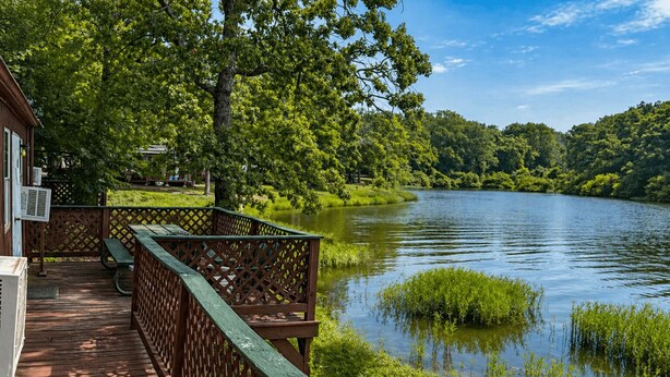 A wooden deck with a lattice railing overlooking a calm lake, surrounded by lush greenery and trees.