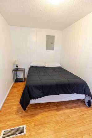 A small, single-bed room with a wooden floor, a bedside table with a lamp, and a wall-mounted light fixture.