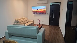 Un salón con un sofá color beige, un sillón verde azulado, una mesa de centro de madera y un televisor en la pared que muestra una escena de atardecer.