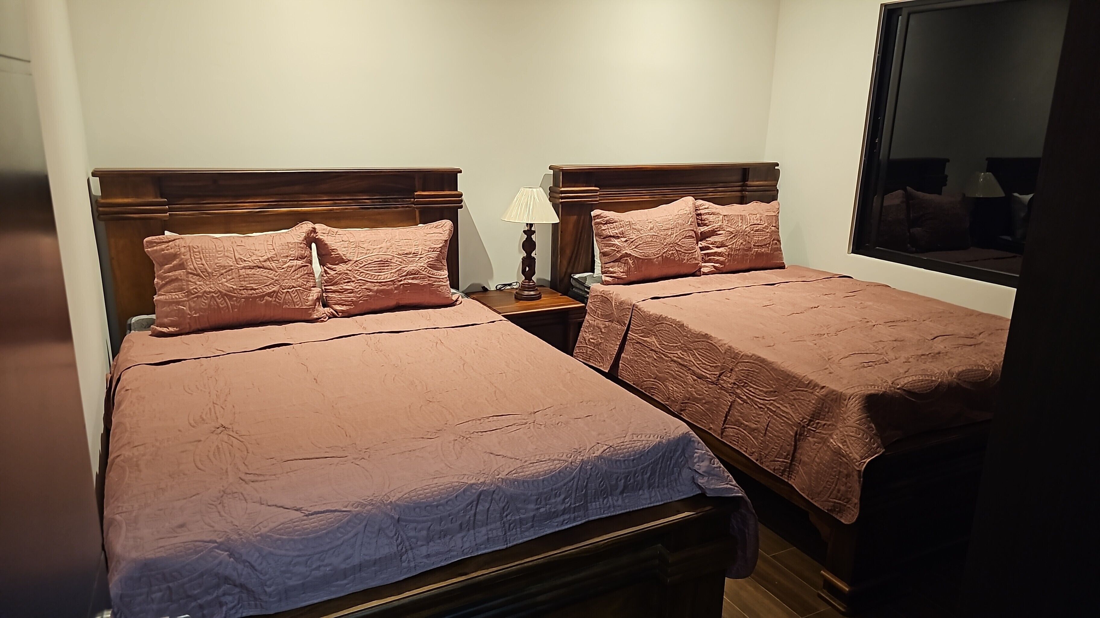 Une chambre avec deux lits, chacun doté d’une tête de lit en bois et d’une couvre-lit rose. Une table de chevet avec une lampe se trouve entre les deux lits.