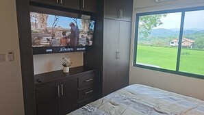 Un dormitorio con cama, televisor montado en un mueble oscuro, y ventana con vistas a un paisaje verde.