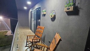 Un pasillo con sillas de madera y una barandilla de metal, una pared oscura con macetas colgantes, y un vidrio al final.