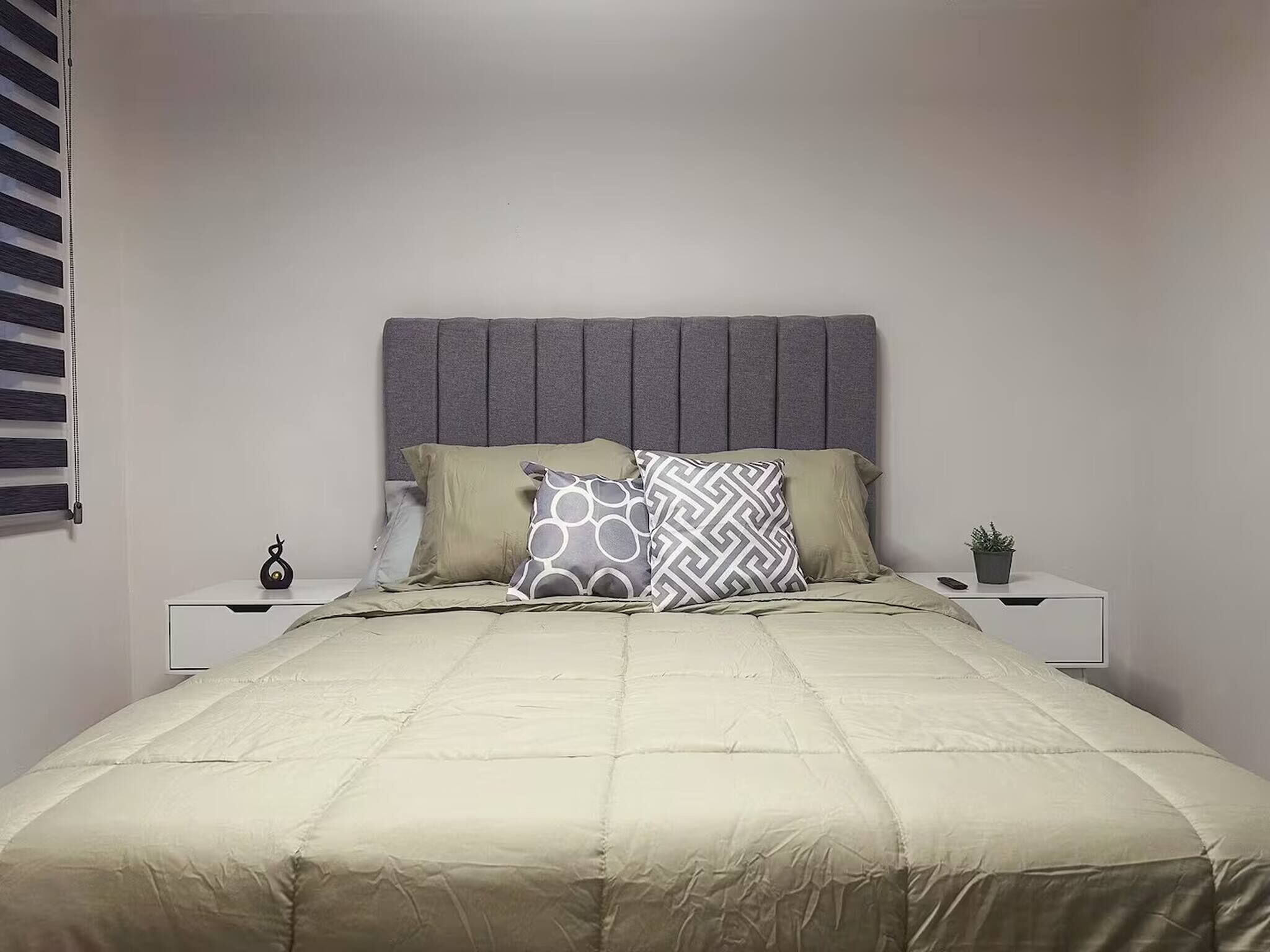 A neatly made bed with a grey headboard, two bedside tables with decorative items, and a small potted plant on the nightstand.