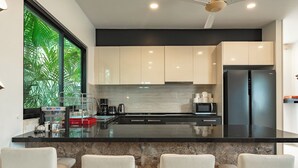 A modern kitchen with a black countertop, white cabinets, and a stainless steel refrigerator. There are three white bar stools and a window with a view of green foliage. - 4Br Near Pier Private Pool & Garden V246 (Rawai)