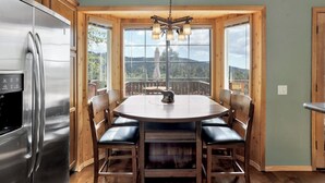 Una cocina con un refrigerador de acero inoxidable, armarios de madera, una mesa redonda con sillas y ventanas amplias que ofrecen vistas al exterior.
