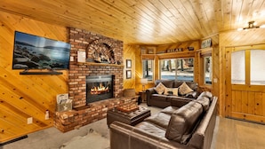 A cozy cabin interior with a fireplace, leather sofa, and a television mounted on the wall.