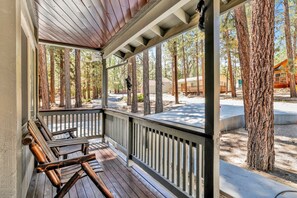 A covered porch with wooden benches, a view of a forested area, and a building in the background.