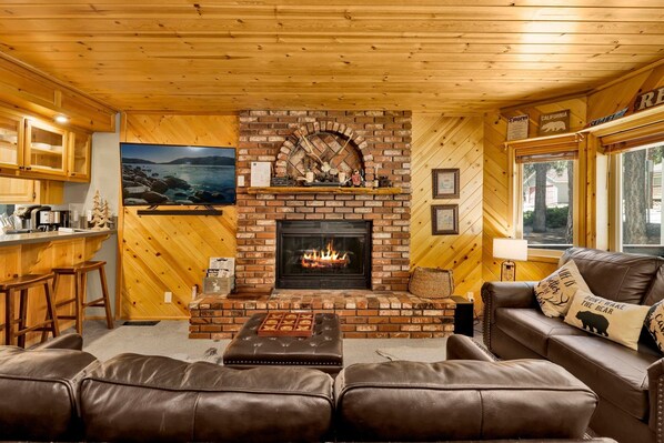 A cozy living room with a fireplace, leather couches, a kitchen area, and a large window.