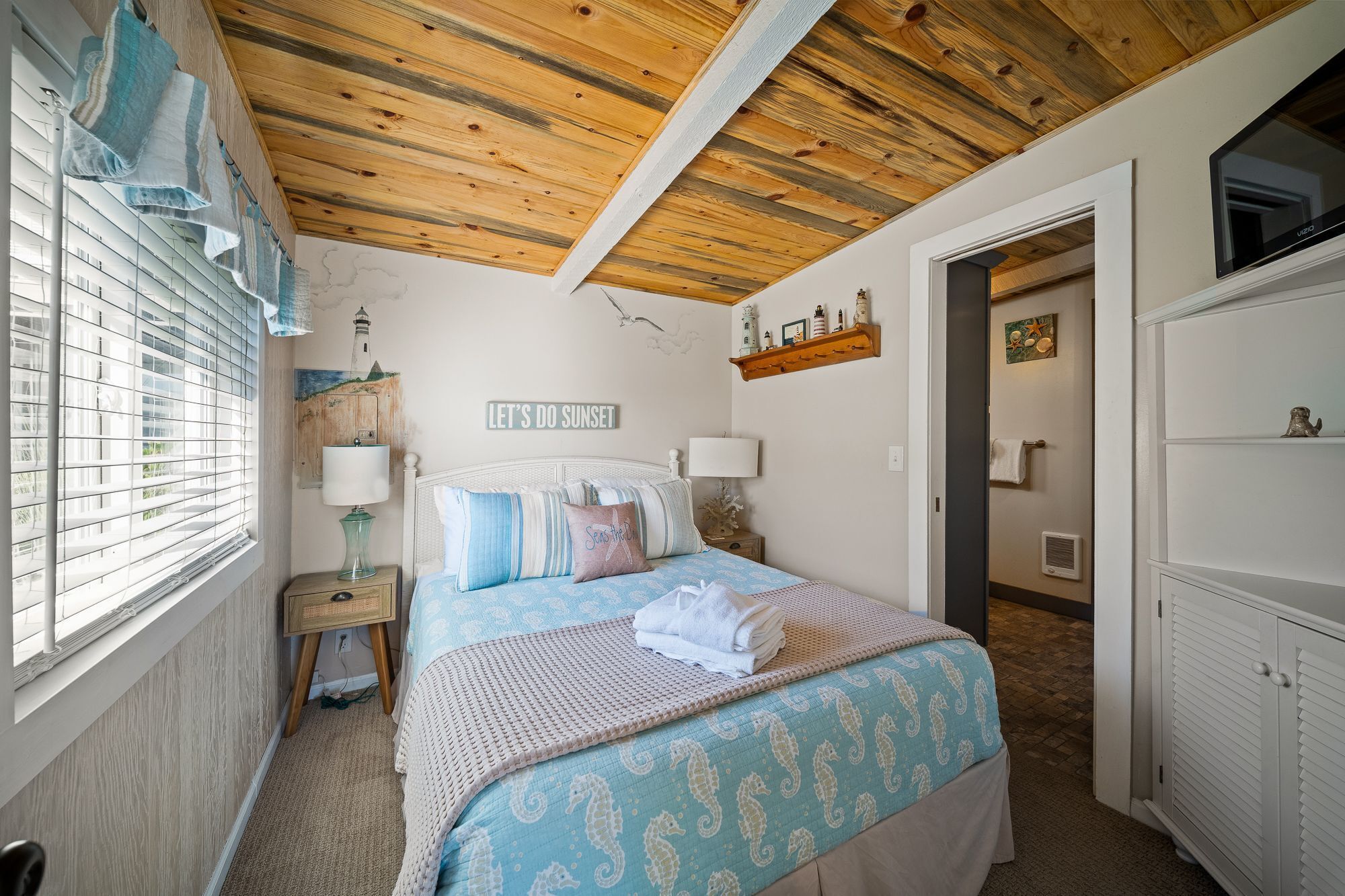 A bedroom with a bed, a nightstand, a wall-mounted shelf with decorative items, and a window with blinds.