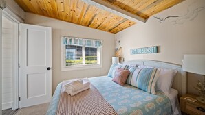 A bedroom with a wooden ceiling, a bed with patterned bedding, a window with blinds, and a sign on the wall. - Cozy Cottage Steps from Beach (Pacific Beach)