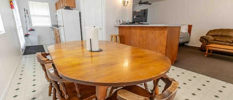 A dining area with a wooden table and chairs, a kitchen in the background, and a living room with a sofa.
