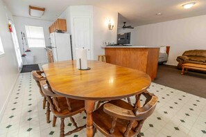 A dining area with a wooden table and chairs, a kitchen in the background, and a living room with a sofa.