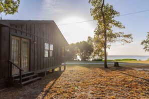 Une cabane rustique avec un porche, une vue sur l’eau et un arbre au premier plan.