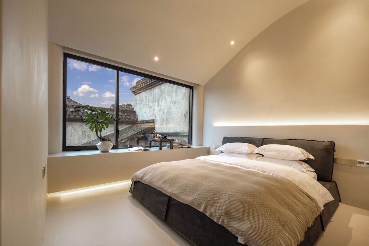 A modern bedroom with a large window, a bed, and a small table with a plant.