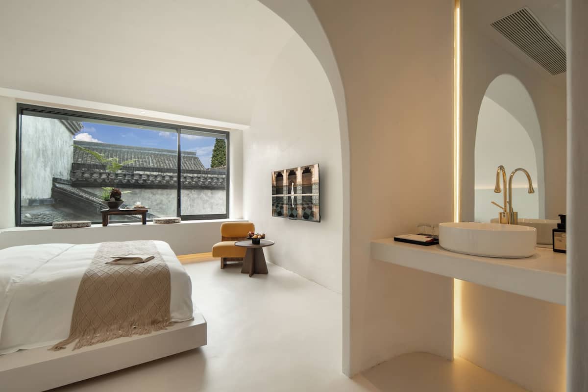 A modern bedroom with a large window, a bed, a bedside table, a chair, and a bathroom with a sink and mirror.