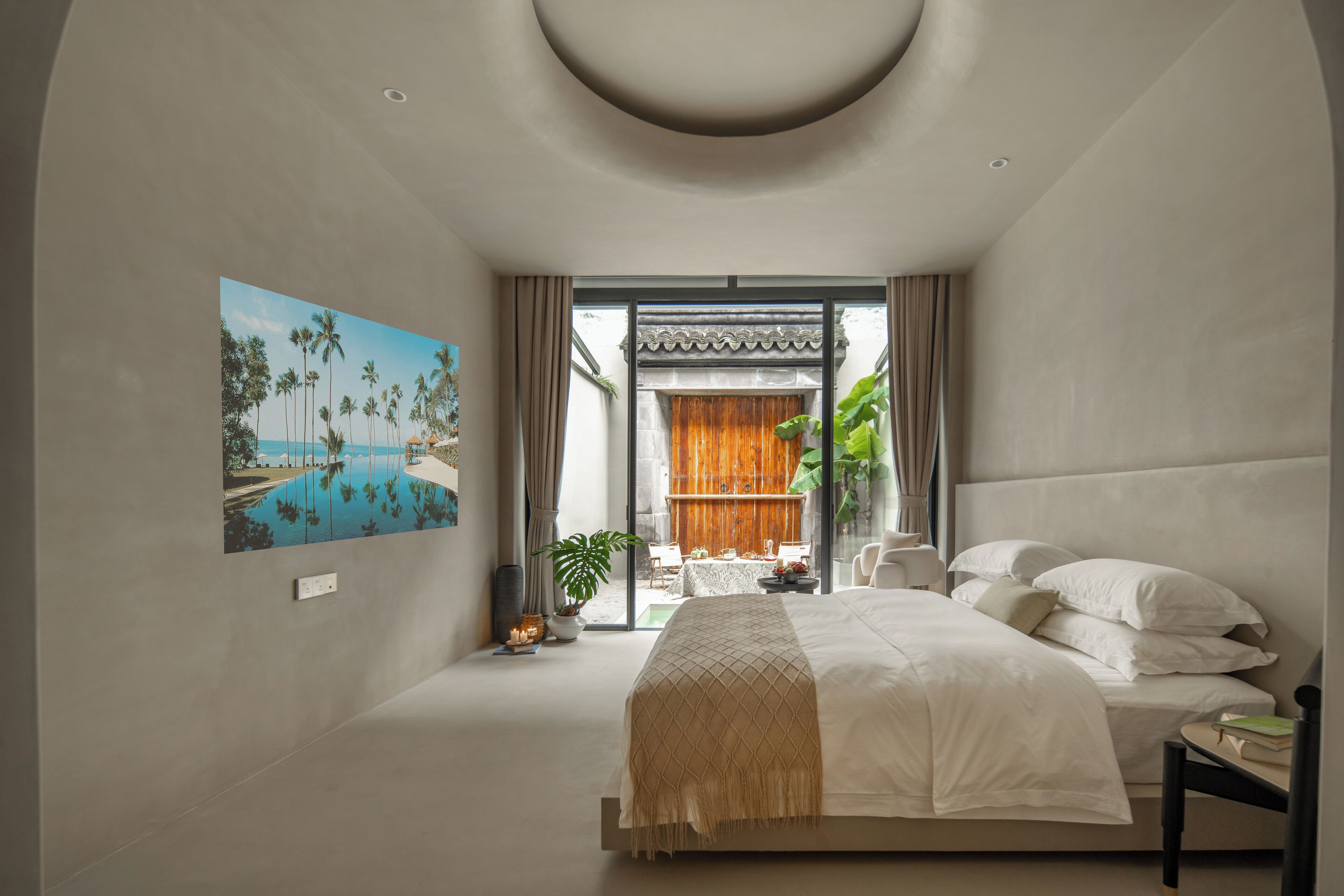 A modern bedroom with a large bed, a view of a tropical outdoor area, and a painting of a beach scene.