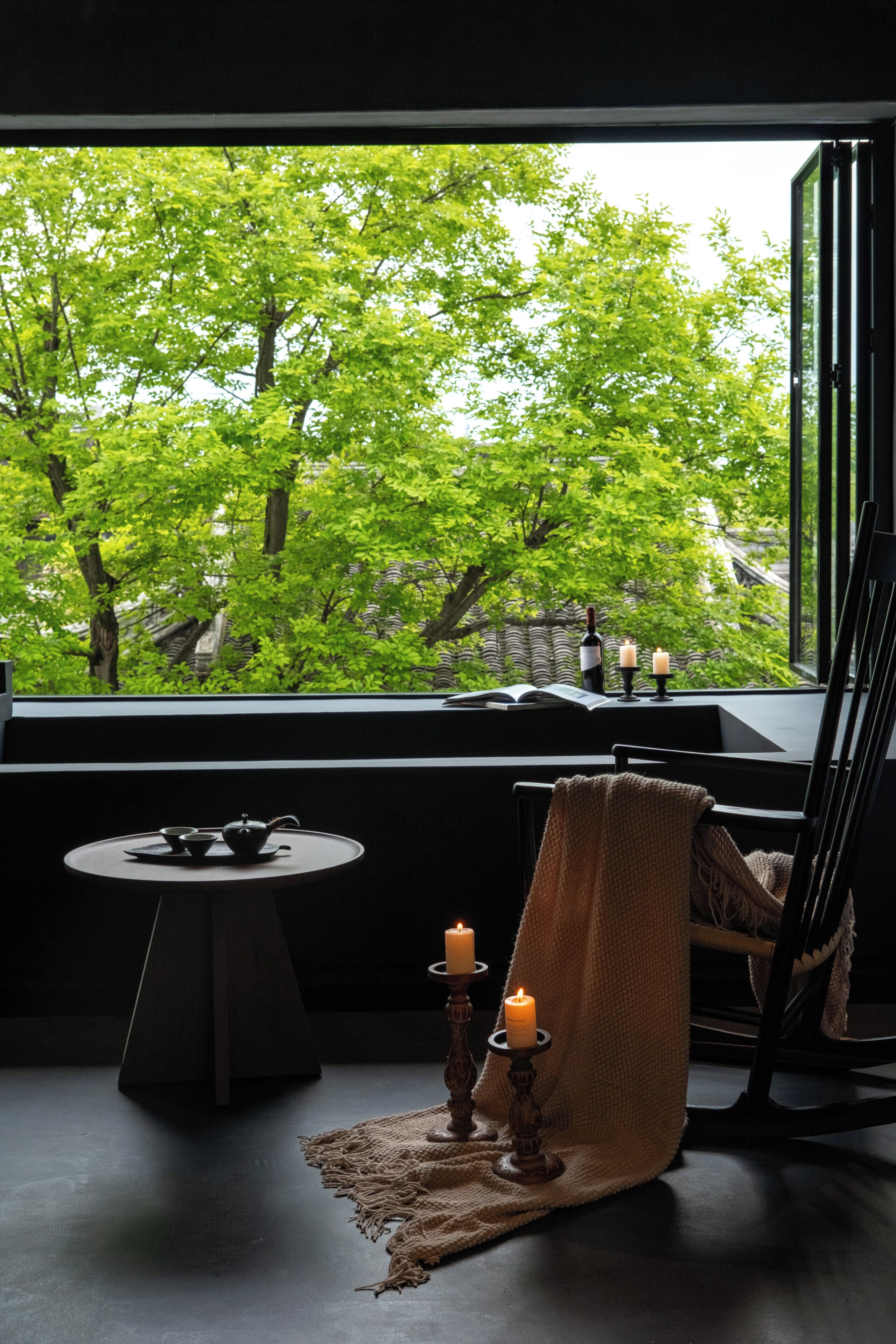 A room with a view of green trees through a window, a small table with a teapot, cups, and a candle, and a chair with a blanket.