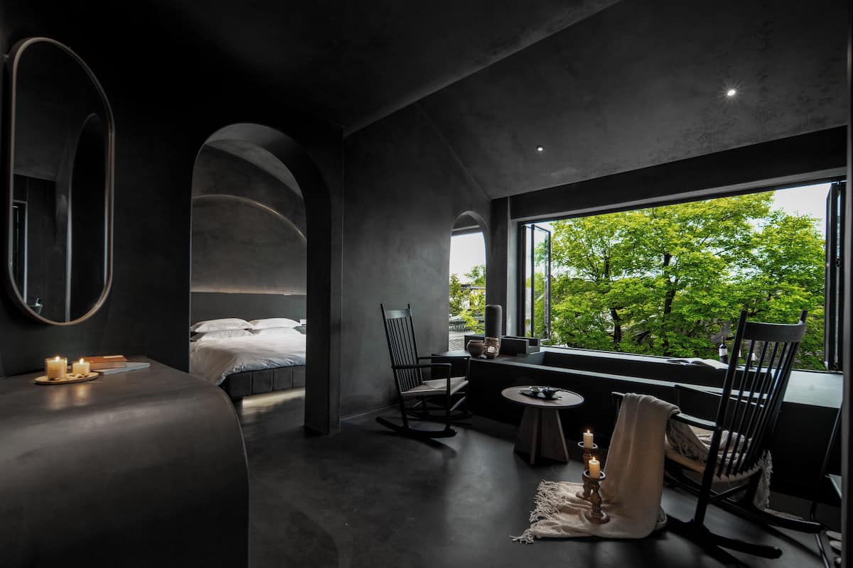 A modern, dark-themed room with a bed, seating area, and a large window offering a view of trees.