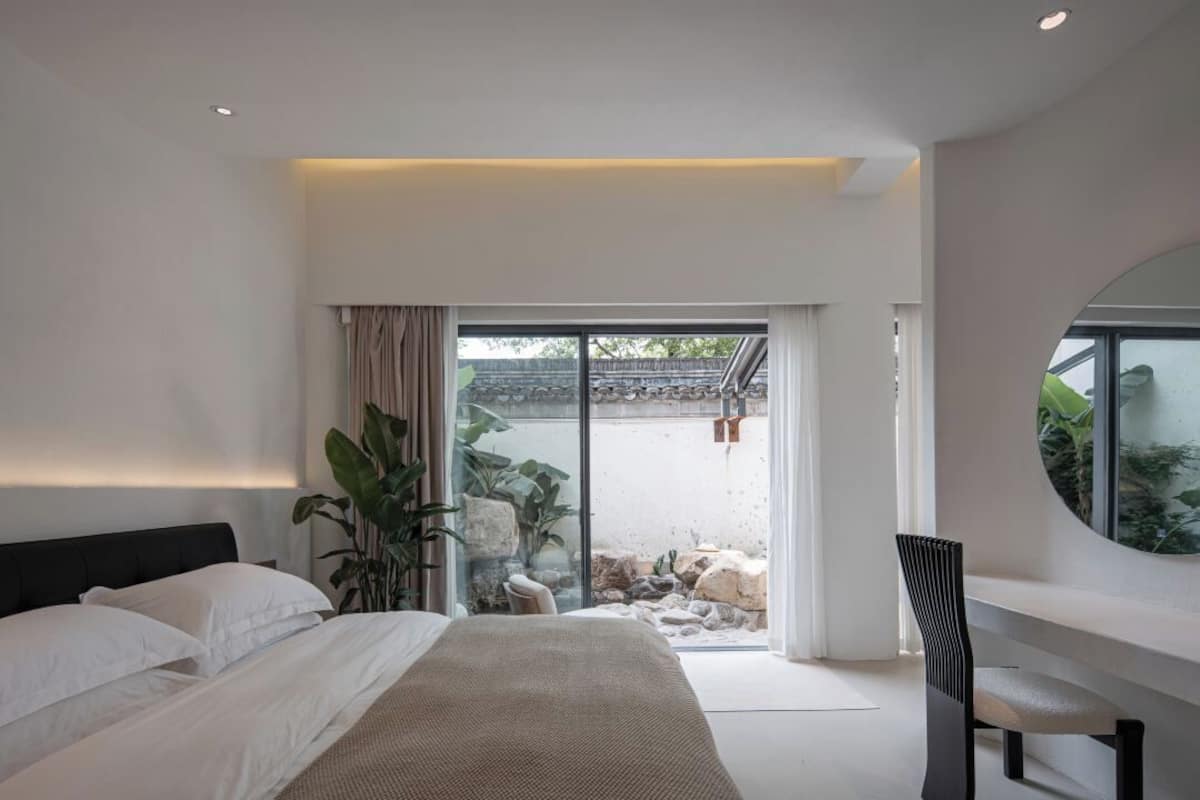 A modern bedroom with a large bed, a seating area, and a view of an outdoor garden.