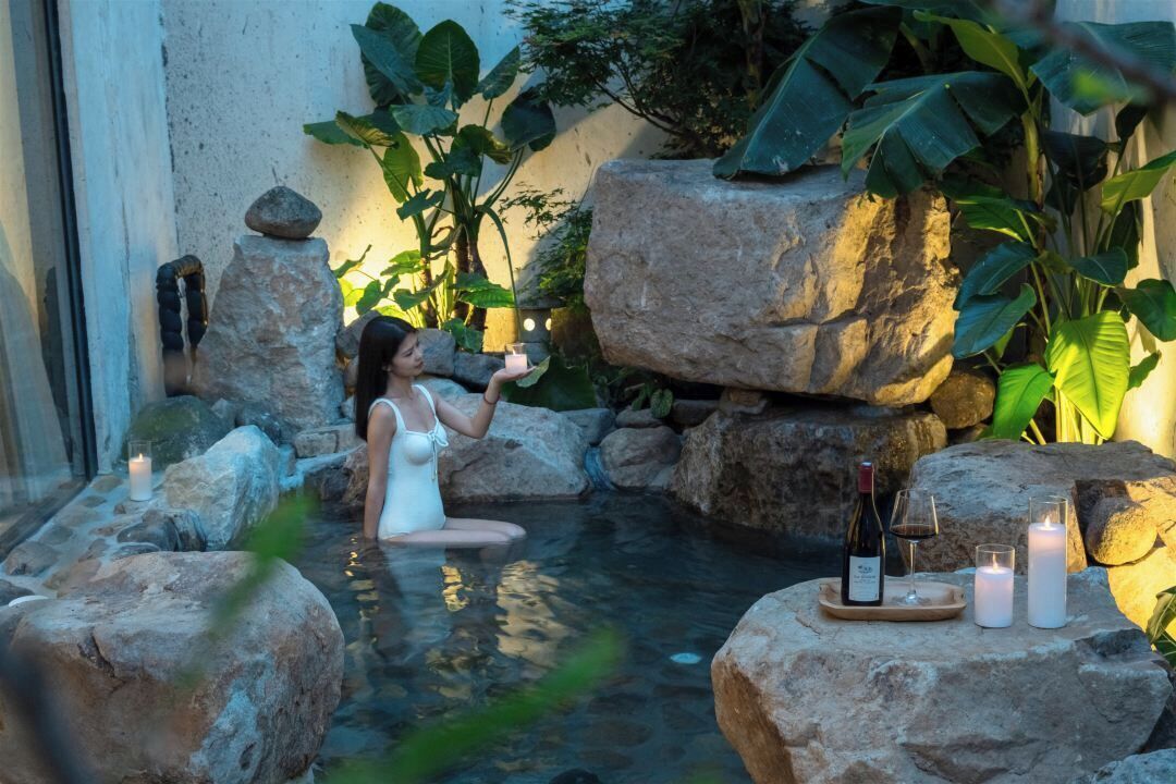 A woman in a white swimsuit sitting in a rock-lined hot tub with candles and a bottle of wine on a tray.