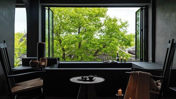 A room with a view of green trees through a large window, a small round table with a teapot, two chairs, and a candle on a side table. - Jiangnanfu Bed and Breakfast (Huzhou)