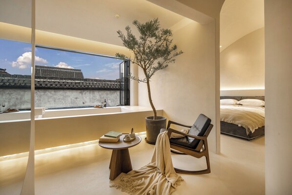 A modern bedroom with a large window, a potted tree, a reading area with books, and a bed with a white duvet.