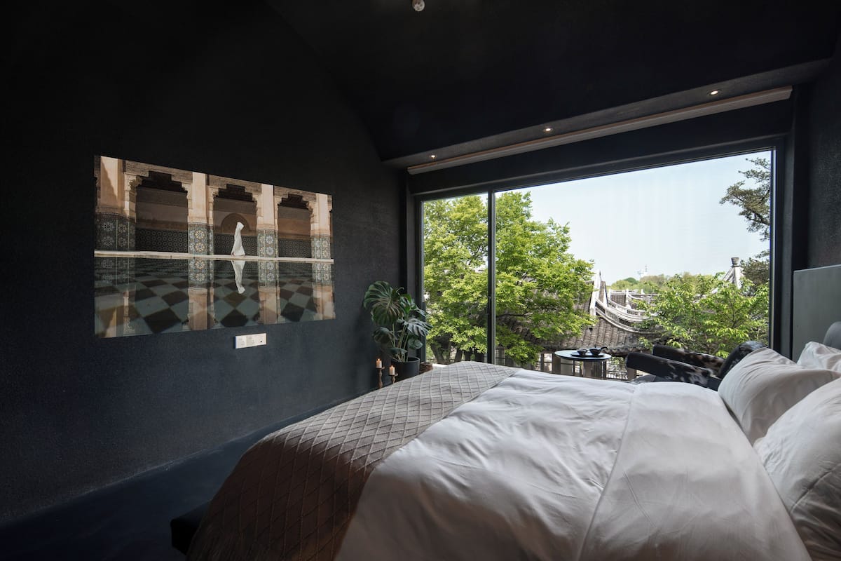 A modern bedroom with a large bed, a view of a traditional courtyard through a large window, and a wall-mounted artwork depicting a historical scene.