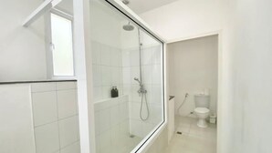 Een moderne badkamer met een douchecabine van glas, witte tegels, een toilet en een klein plankje met toiletartikelen.