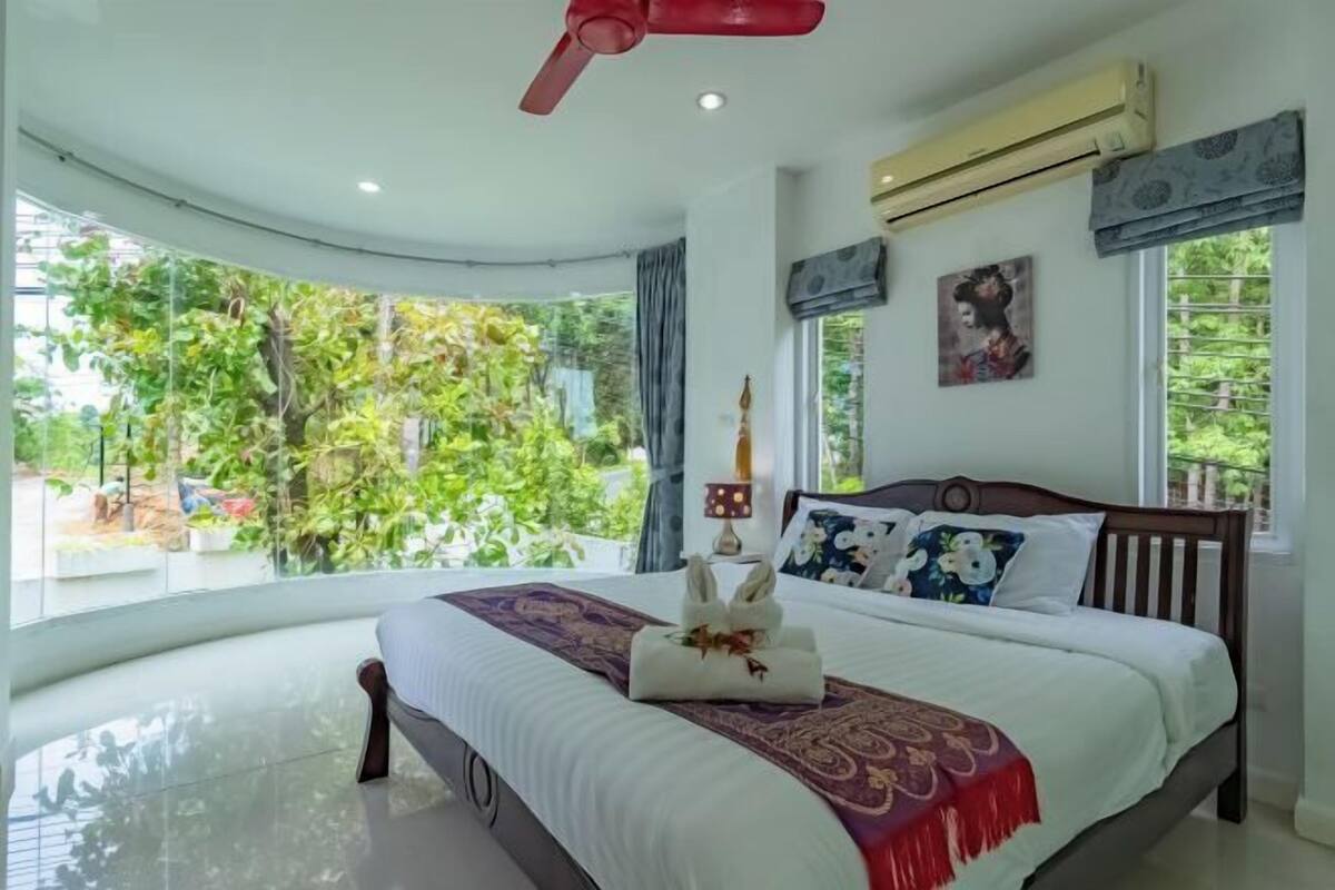 Une chambre à coucher comprenant un lit, un ventilateur de plafond, un appareil de climatisation, une fenêtre donnant sur un espace verdoyant et un luminaire fixé au mur.