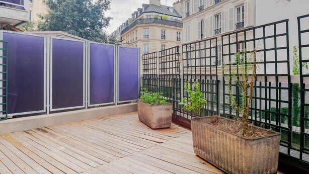 Une terrasse en bois avec une clôture en métal, des plantes en pot et une vue sur les bâtiments en arrière-plan.
