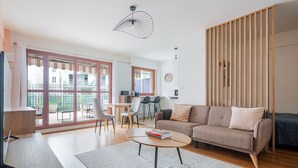 Un salon moderne avec un canapé, une table basse et des fauteuils. On y trouve également un coin repas avec une table et des chaises, et une cuisine à l’arrière-plan.