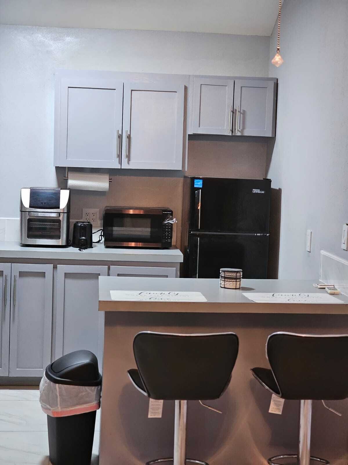 Cocina compacta con microondas, tostadora y refrigerador. Hay tres taburetes de bar y un cubo de basura.