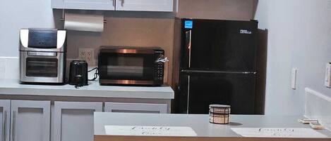 Cocina compacta con microondas, tostadora y refrigerador. Hay tres taburetes de bar y un cubo de basura.