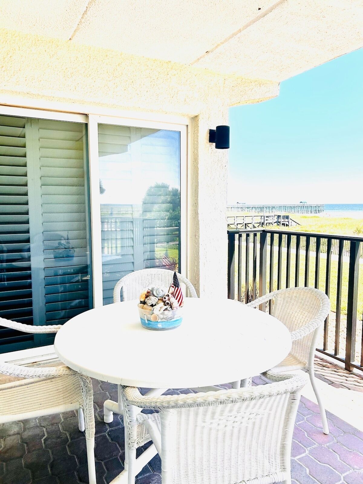 A round white table with wicker chairs on a balcony with a view of the beach and a wooden pier.