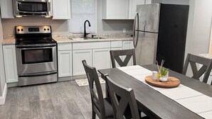 Una cocina moderna con electrodomésticos de acero inoxidable, una zona de comedor con una mesa y sillas de madera, y un piso gris.