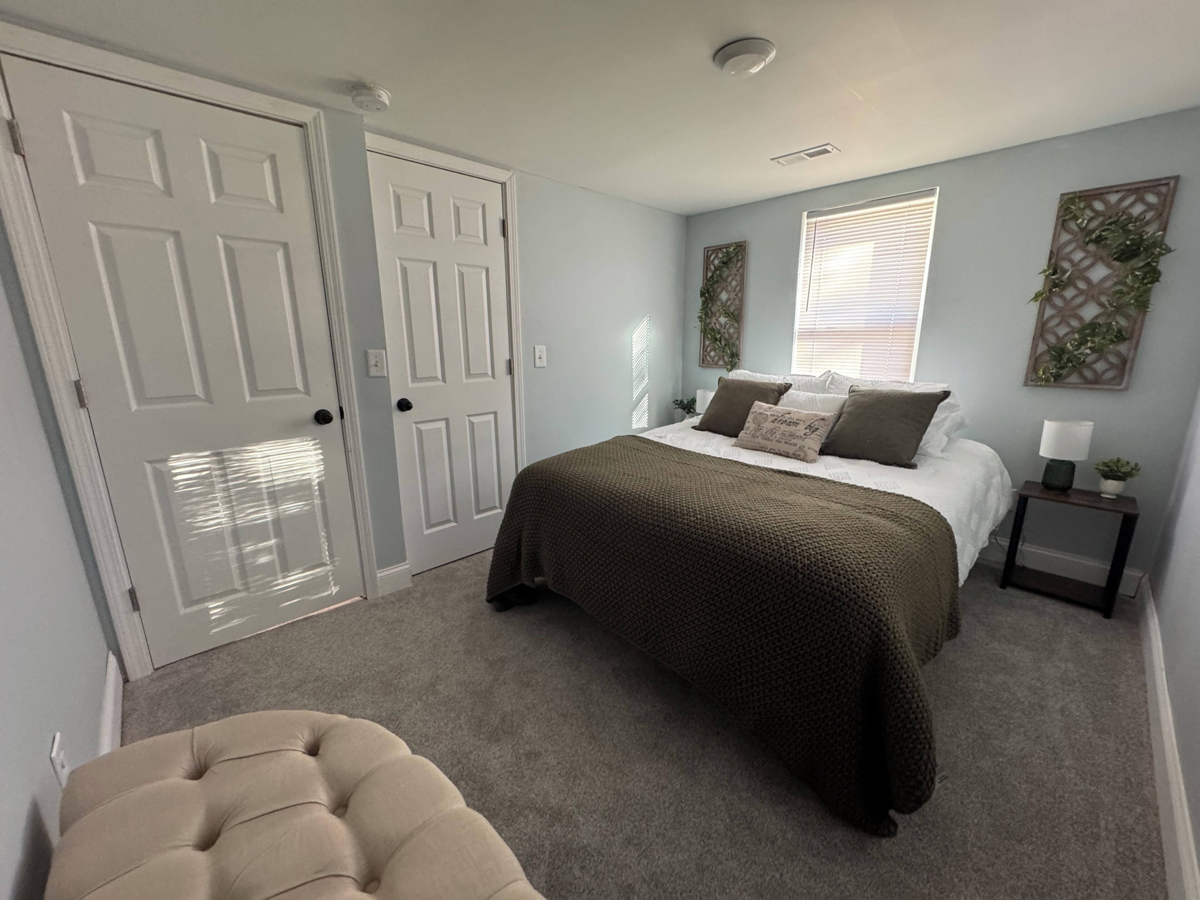 Une chambre à coucher comprenant un lit, deux portes, une fenêtre avec des stores, une table de chevet avec une lampe et un élément décoratif mural.