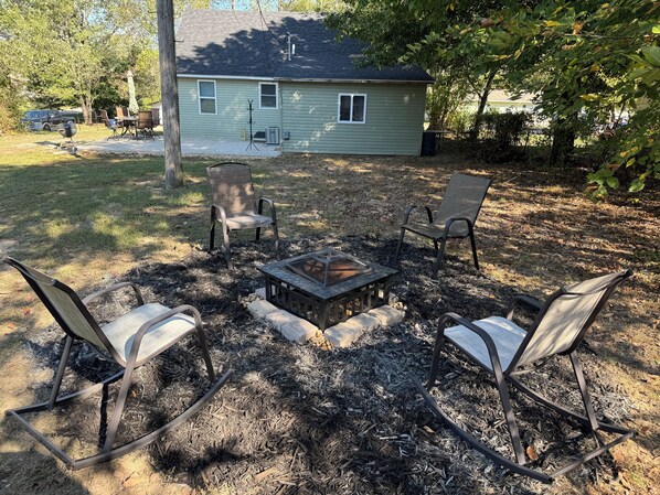 A backyard with a fire pit surrounded by four chairs, a covered table, and a house in the background.