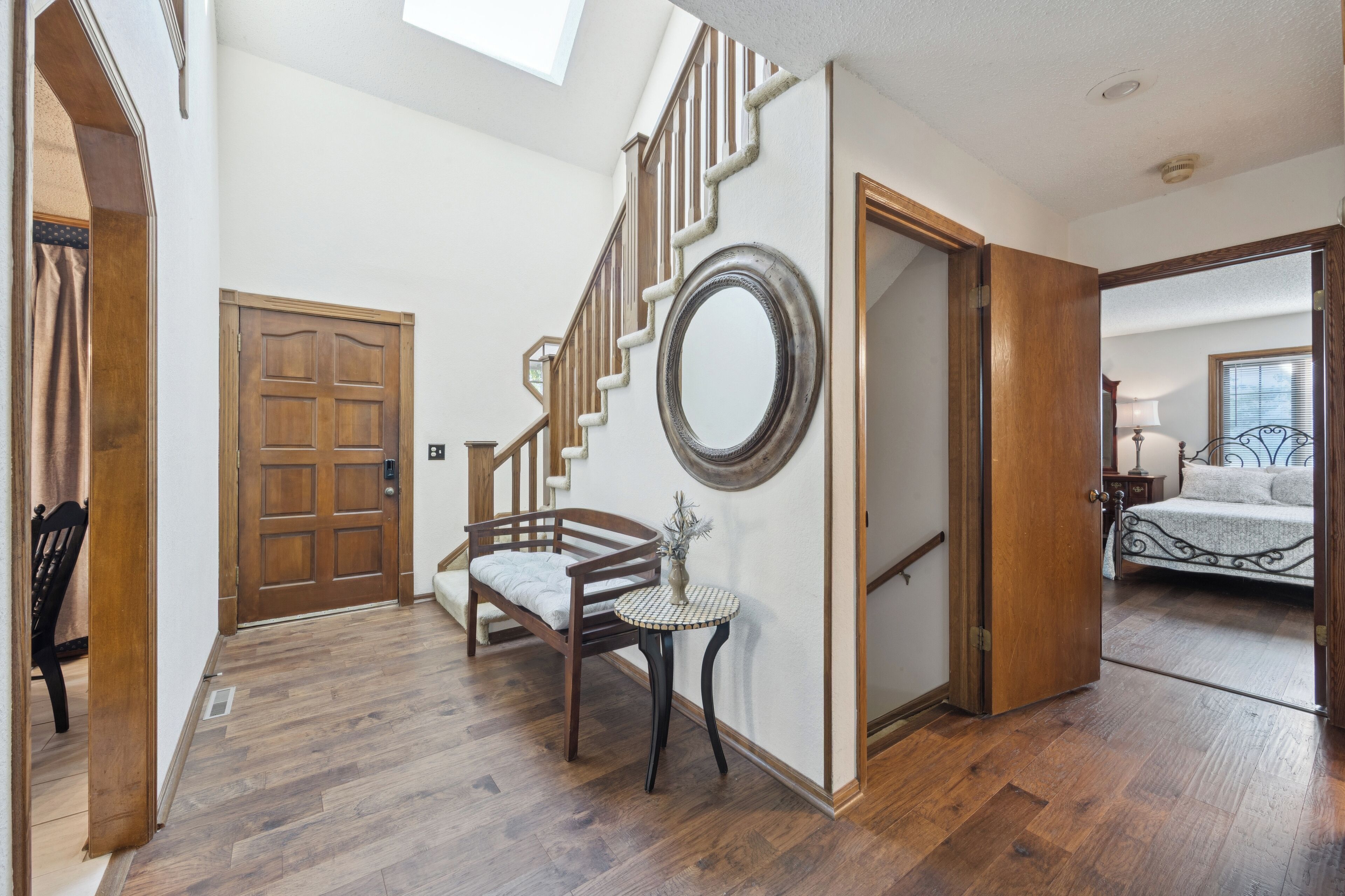 A spacious hallway with a wooden staircase, a round mirror on the wall, and a bed in the adjacent room.