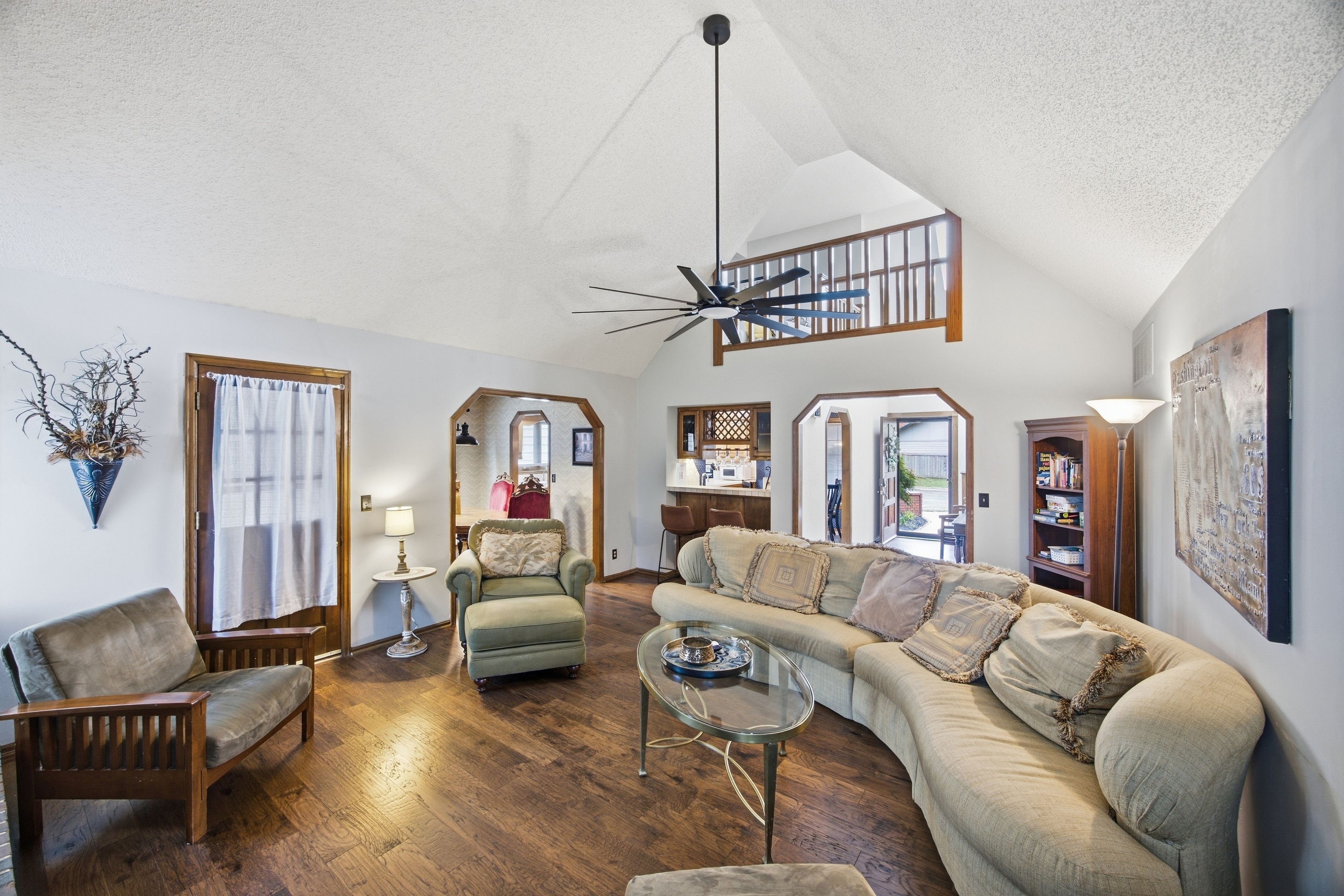 A spacious living room with a high ceiling, a ceiling fan, a wooden floor, a sofa, a chair, a coffee table, and a bookshelf.