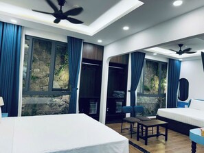 A modern hotel room with a large bed, a desk, and a chair. There are windows with curtains, a ceiling fan, and a stone wall visible through the windows.