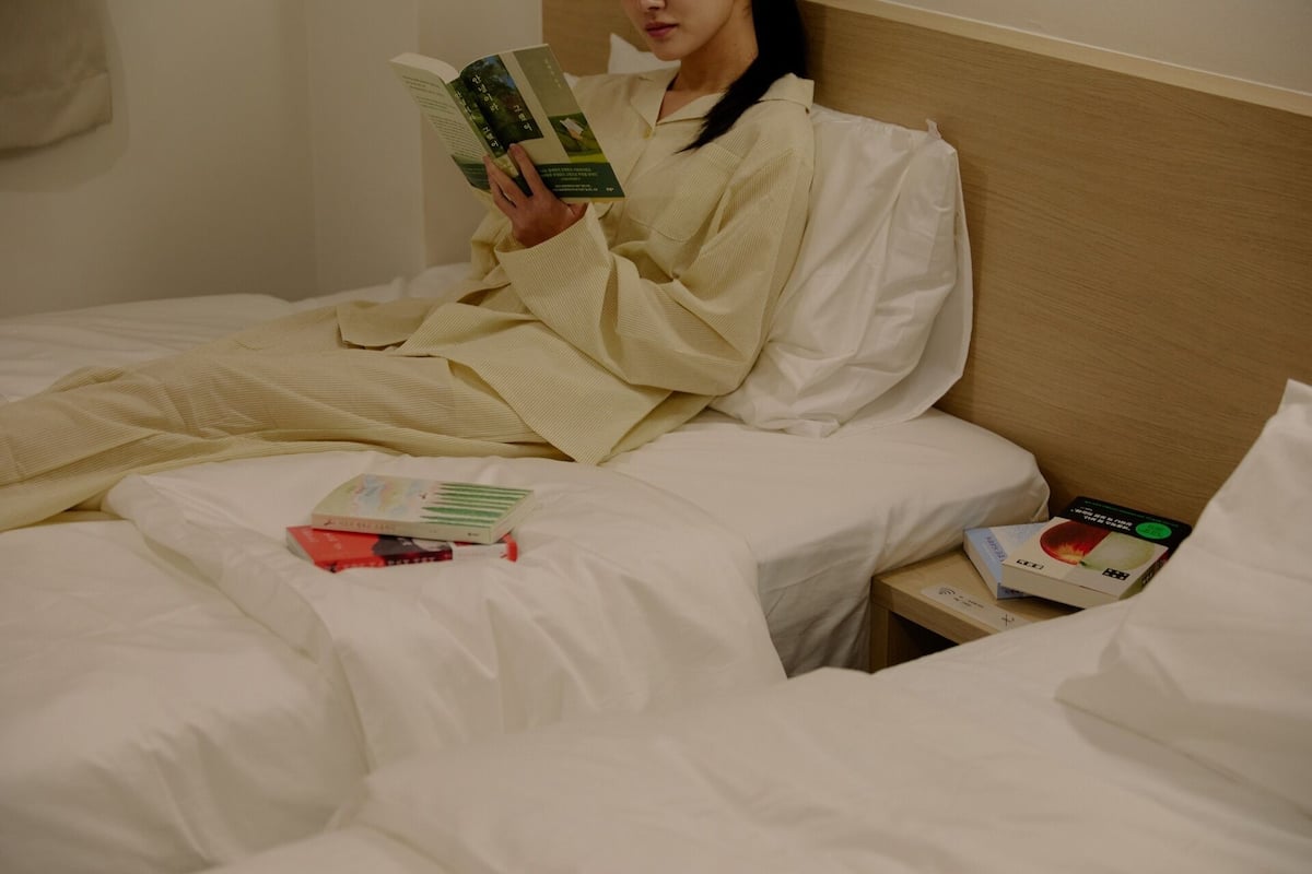 Eine Person in einem Hotelzimmer, die ein Buch im Bett mit weißer Bettwäsche liest. Auf dem Bett liegen zwei Bücher und ein kleiner Nachttisch mit einem Buch und einer Schachtel.