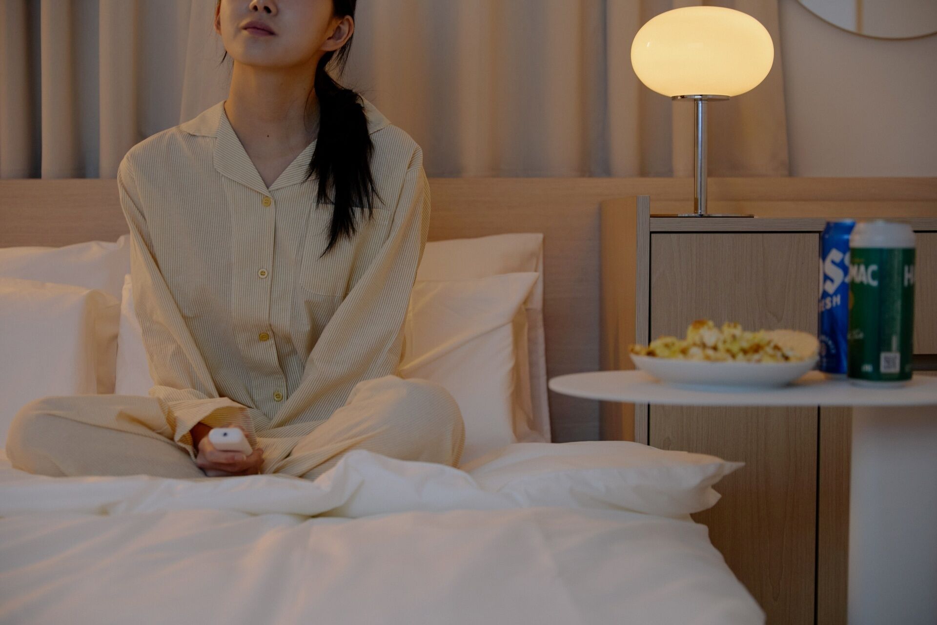A person sitting on a bed in a hotel room with a lamp and snacks on a small table.