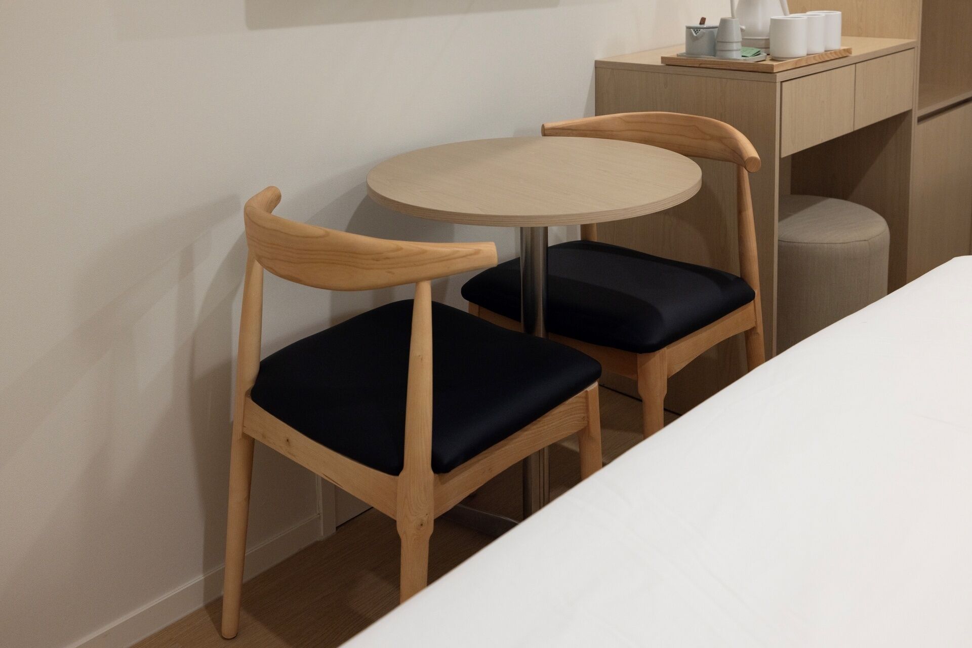A small dining area with two wooden chairs with dark cushions, a round table, and a wooden cabinet with a tray holding cups and a kettle.