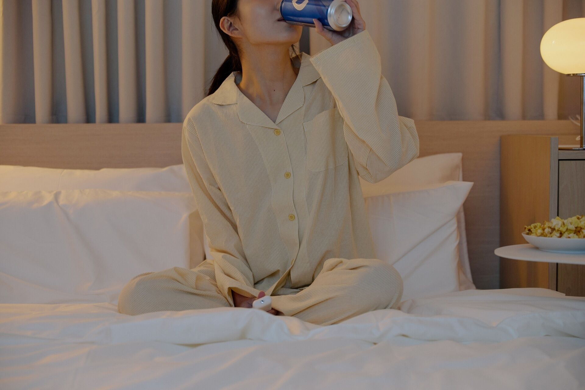 Una persona sentada en una cama en una habitación con cabecera, una mesita de noche con lámpara y un tazón de cereal.