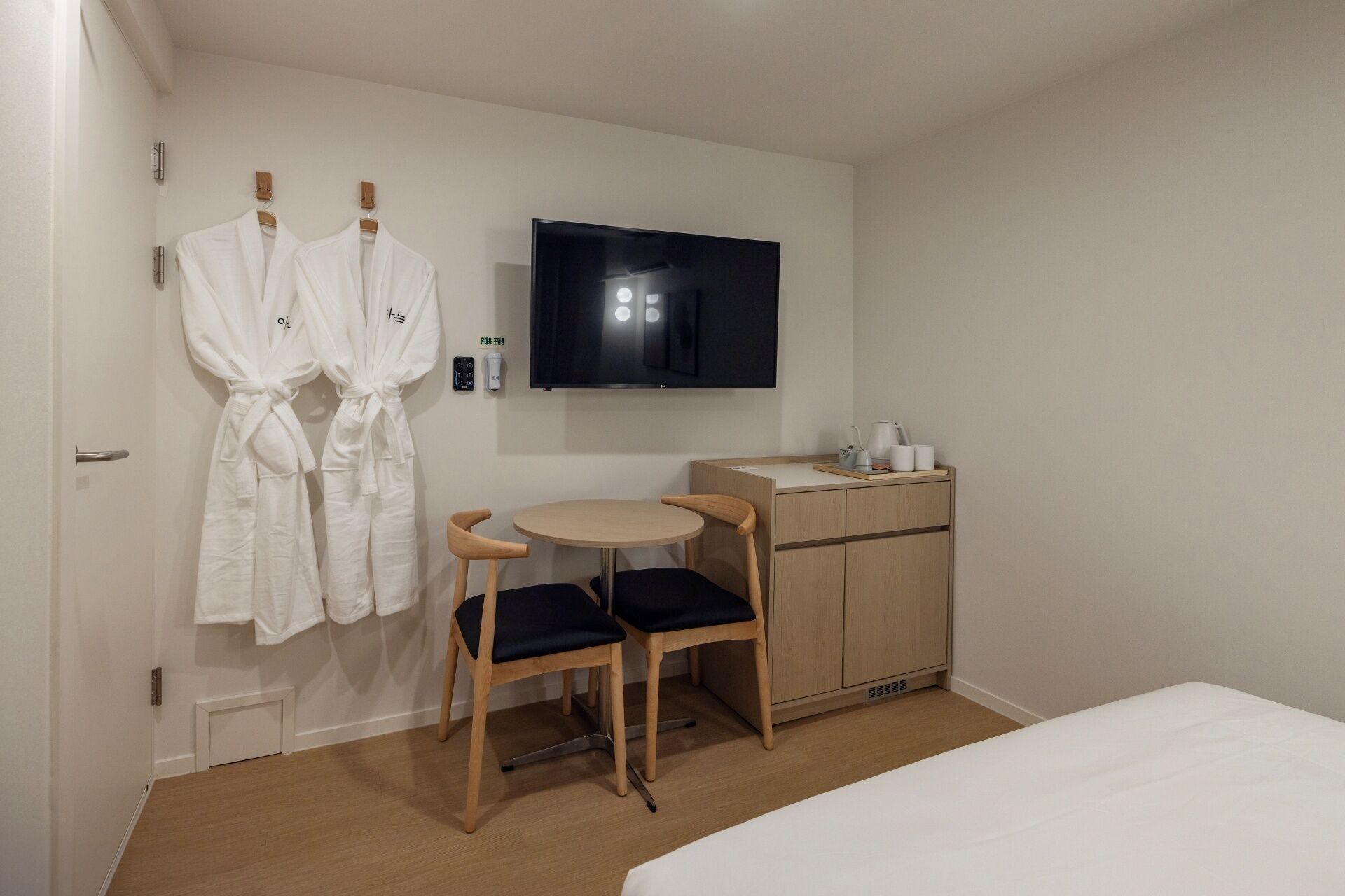 Habitación de hotel con cama, escritorio con silla, televisor en la pared y dos batas blancas colgadas en una puerta.