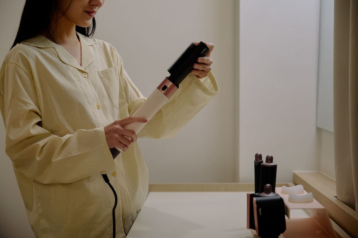 Eine Person in einem gelbgestreiften Bademantel hält eine schwarze Haarbürste und ein weißes Objekt, stehend in einem Raum mit einem Holztresen und einem Spiegel.
